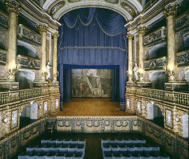 Auditorium mit blauen Stühlen, einem blauen Vorhang, kunstvoll gestalteten Wänden und Skulpturen sowie einer geschnitzten Decke, das eine hell erleuchtete Bühne präsentiert.