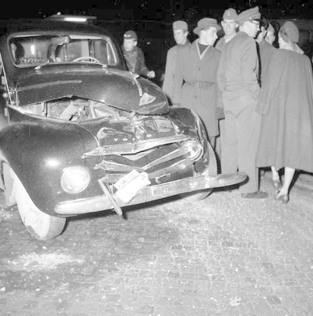 Schwarz-weiß-Bild eines schwer beschädigten Autos am Straßenrand mit einer Gruppe von Menschen, die es inspizieren.