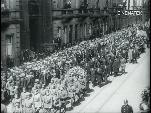 Schwarzes und weißes Foto einer großen Menge von Menschen, die in einer Parade marschieren, einige halten Gewehre und gehen an einem Gebäude vorbei, mit einem Wasserzeichen oben rechts.