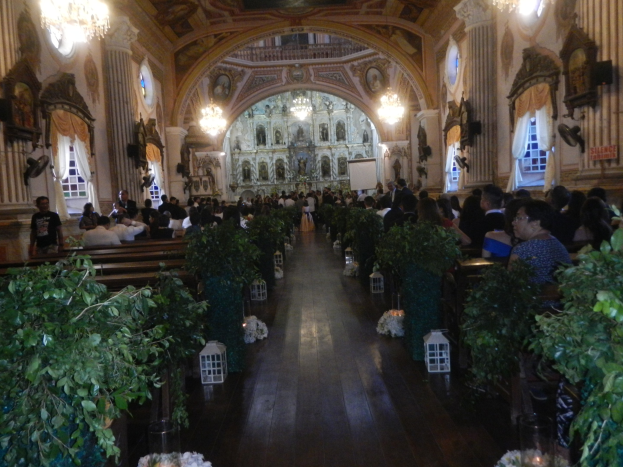 Eine Trauung in einer Kirche mit Gästen auf Bänken sitzend und im Gang stehend, dekoriert mit Zimmerpflanzen, Skulpturen an den Wänden, Vorhängen an den Fenstern und hängenden Kronleuchtern, mit einem Bildschirm im Hintergrund.