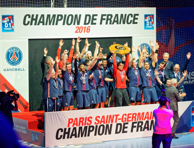 Gruppe von Männern auf einer Bühne, die einen Pokal halten, mit einer Person, die filmt, und einem "Champion de France 2016"-Schild im Hintergrund.
