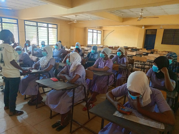 Eine Klasse in Nigeria mit Schülern, die Gesichtsmasken tragen und auf Stühlen und Stehplätzen sitzen, umgeben von Büchern und Gegenständen, mit Fenstern, einer Tür, Lampen und Ventilatoren im Hintergrund.