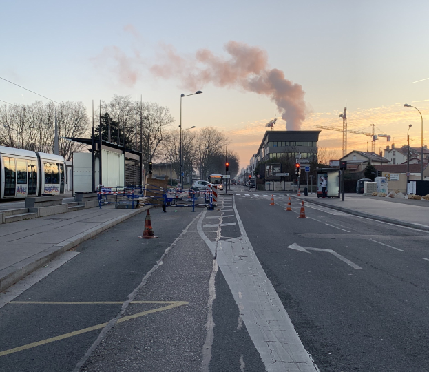 Eine Stadtstraße mit Absperrgittern am Rand, ein Bus auf der linken Seite, Laternenpfähle, Bäume, Gebäude, Kräne im Hintergrund und Rauch, der aus einem entfernten Gebäude aufsteigt, unter einem Himmel.
