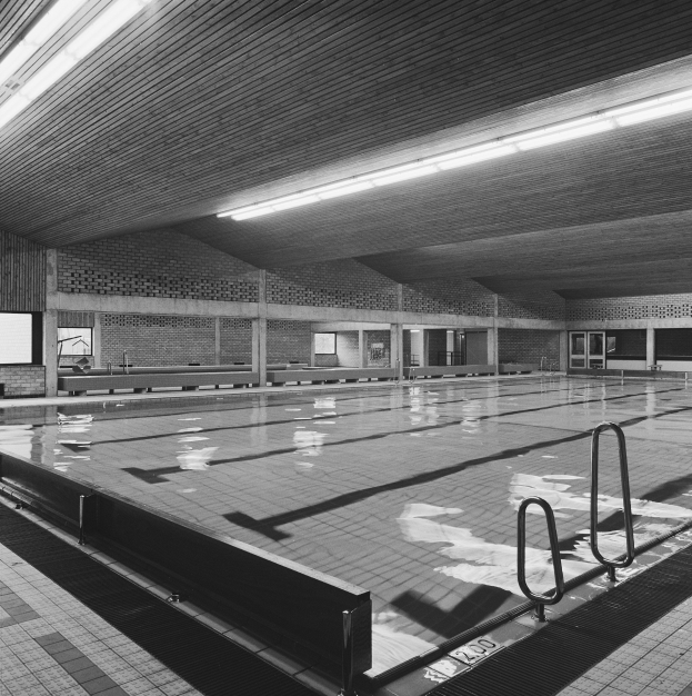 Schwarz-weiß-Foto eines Hallenbads in einem großen Gebäude mit gefliestem Boden, Metallstreben, Wänden, Fenstern und Deckenleuchten.