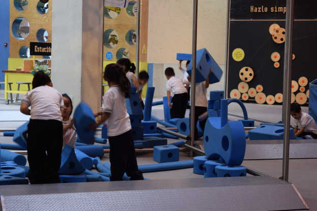 Kinder beim Spielen in einer Turnhalle mit Gegenständen in den Händen und einer Texttafel an der Wand sowie Stangen auf der rechten Seite.