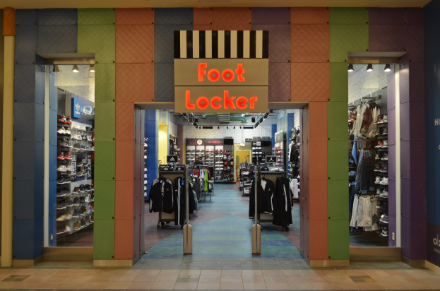 Innenansicht eines Foot Locker-Schuhgeschäfts mit Kleidung an Bügeln, Waren auf Gestellen und Deckenbeleuchtung.