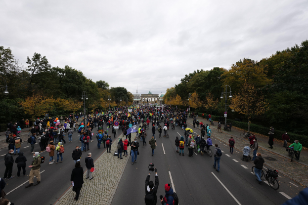 Eine große Gruppe von Menschen marschiert auf einer von Bäumen gesäumten Straße mit Laternenpfählen, einem Gebäude im Hintergrund und einem klaren Himmel, einige halten Kameras während einer Demonstration in Berlin.