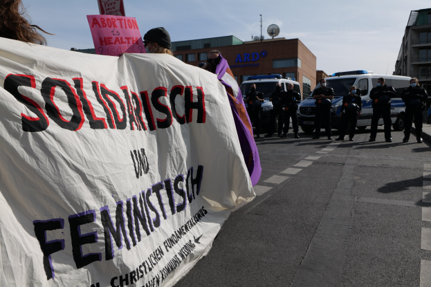 Gruppe von Menschen, die mit einem "Solidarität und Feminismus"-Schild die Straße entlangmarschieren, mit parkenden Fahrzeugen und Gebäuden im Hintergrund unter einem klaren blauen Himmel.