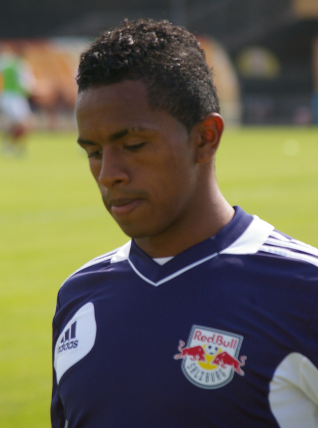Junger Mann in blauem T-Shirt auf einem Fußballfeld, der wie ein Flügelstürmer mit kurzen dunklen Haaren und entschlossener Miene aussieht.