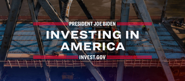 Präsident Joe Biden neben einer Brücke mit dem Text "In America investieren" und verstreuten Gegenständen im Vordergrund.