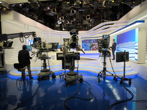 Ein TV-Studio mit mehreren Kameras auf dem Boden, einer sitzenden Person im Vordergrund und einigen stehenden Personen im Hintergrund, mit an der Decke montierten Lichtern, die auf eine Livesendung hinweisen.