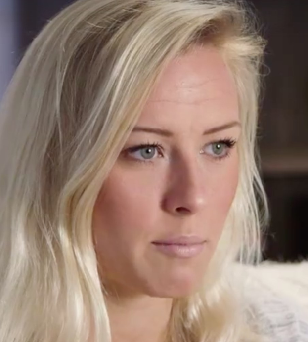 Eine Frau mit langen blonden Haaren und blauen Augen im Vordergrund, die besorgt wirkt, mit einem unscharfen Hintergrund.