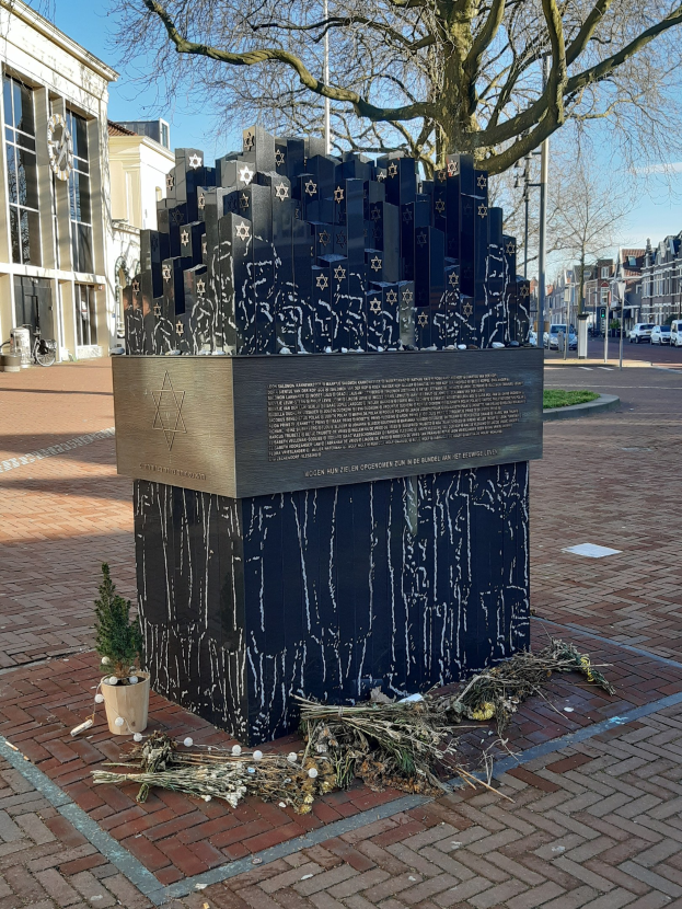 Denkmal in einem Stadtplatz für Holocaust-Opfer mit einem Baum davor, einem Topf mit einer Pflanze, verstreuten trockenen Blättern und Gebäuden, Fahrzeugen, Pfählen und Bäumen im Hintergrund.